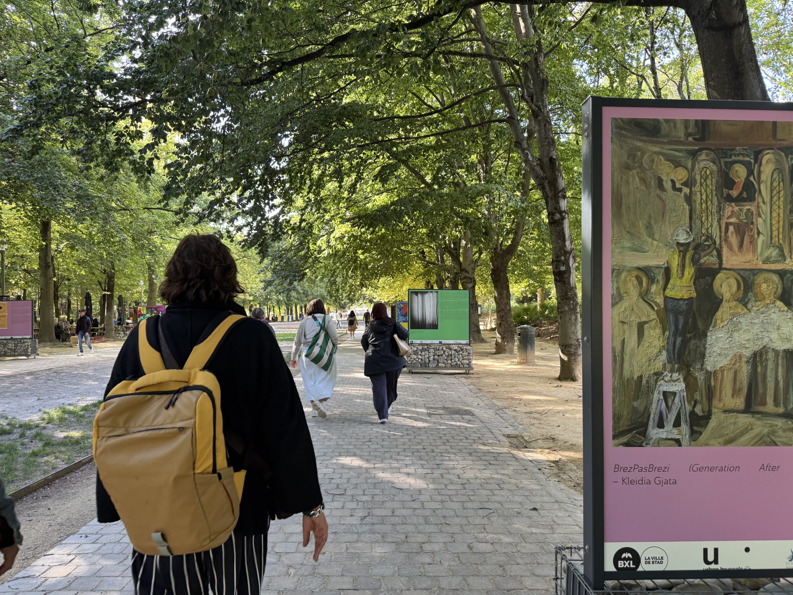 Visitor waling through an outdoor exhibition in a Brussels park, with artworks from the Living Without Leaving campaign displayed on panels along a tree-lined alley.